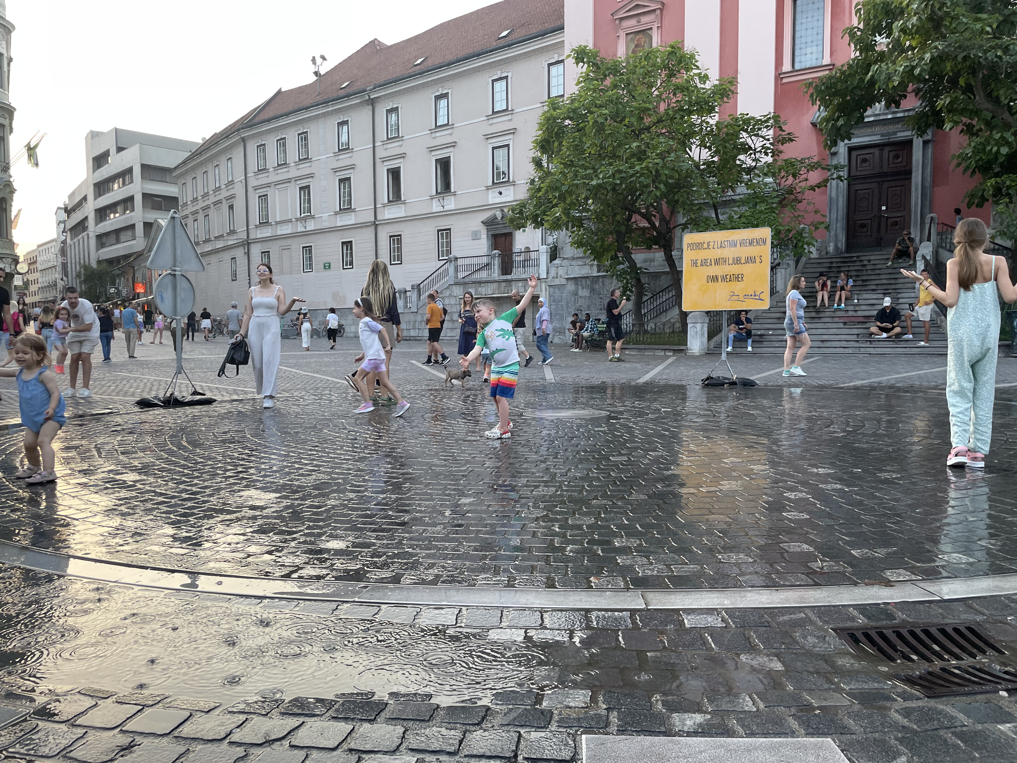 A group of people playing in an apparent rain shower in a pedestrian square. László is centered with his arms out.