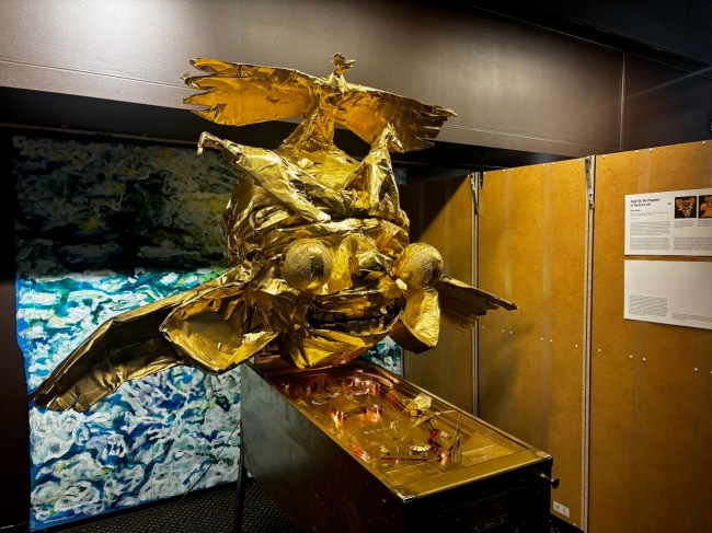 A golden creature made from duct tape and foam with an eagle on its head, large ears, wings, and bug eyes, mounted on top of a gold-painted pinball machine.